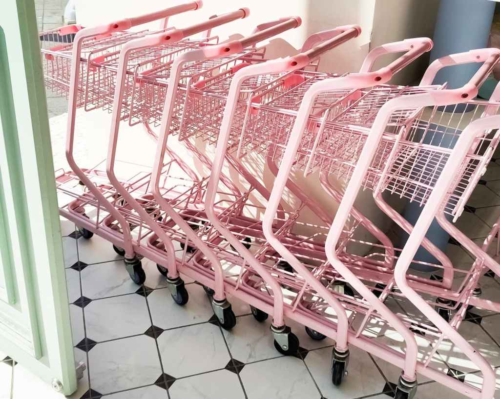 一排粉色購物推車在日光下整齊排列，以日常消費場景呼應「材料換了就能永續嗎」的提問，暗示永續思考不只停在材料選擇。